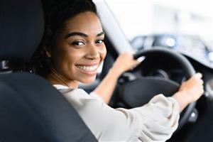 A women smiles from the front seat of a car, her hands are on the steering wheel.