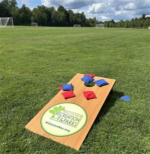 A cornhole board lays in the grass on a sunny day, showing Williston Recreations logo on the front of the board.